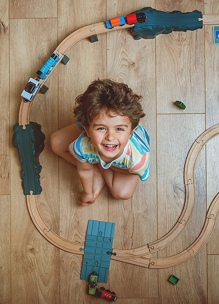 Smiling child sits inside a wooden toy train track on a wooden floor